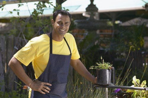 A smiling man wearing a yellow T-shirt and dark apron stands in a garden, holding a potted plant with green foliage and purple flowers on a metallic plant stand. The background features a lush garden setting with mature trees, shrubs, and flowering plants, along with a wooden fence partially visible. The garden has a mix of grass and flower beds, with a paved area or patio nearby, and sunlight filtering through the trees, indicating a bright, clear day. This outdoor space appears well-maintained, suitable for gardening and landscaping activities, reflecting typical features of a residential garden in Lewisham. The scene emphasizes gardening care and outdoor aesthetic enhancement consistent with services offered by Garden Maintenance Lewisham.